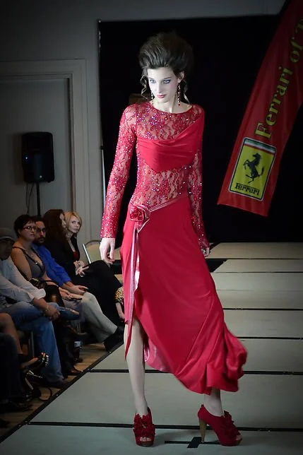 Model walking down runway in a red lace dress with long sleeves, floral embellishments, and a slit, at a fashion show with audience and Ferrari banner in background.