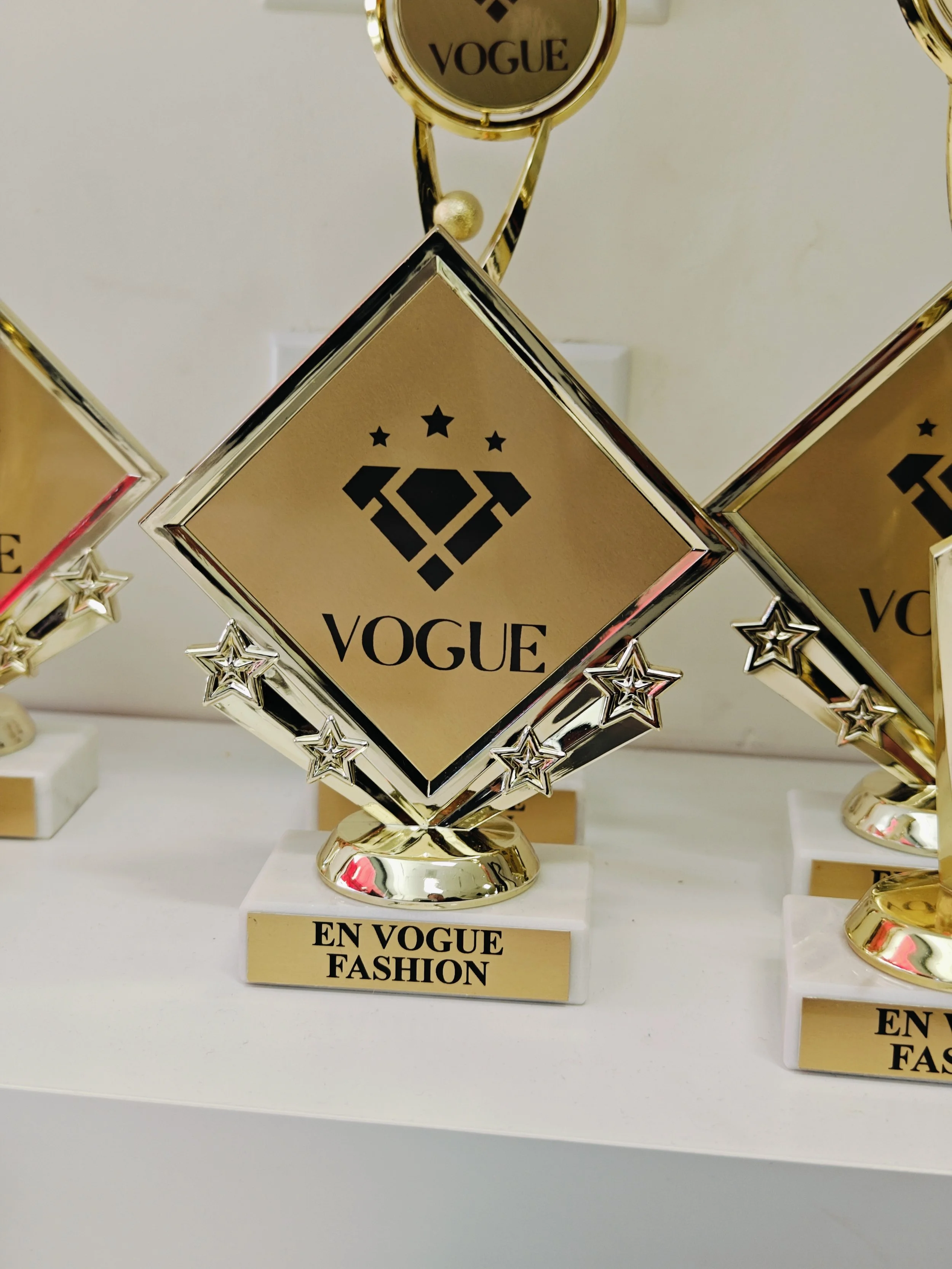 Gold and silver trophies with the word 'VOGUE' and a logo, displayed on a shelf with a label 'EN VOGUE FASHION'.