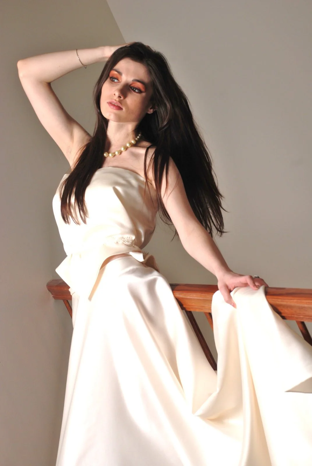 A woman with long dark hair and makeup wearing a cream satin dress with a pearl necklace, posing on a staircase.