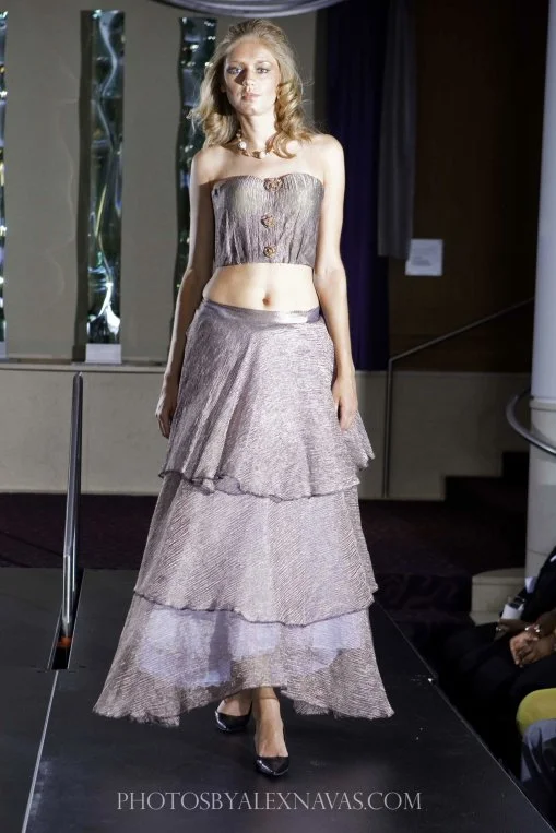 A woman walking on a fashion runway, wearing a strapless metallic top and a tiered, metallic maxi skirt with high heels.
