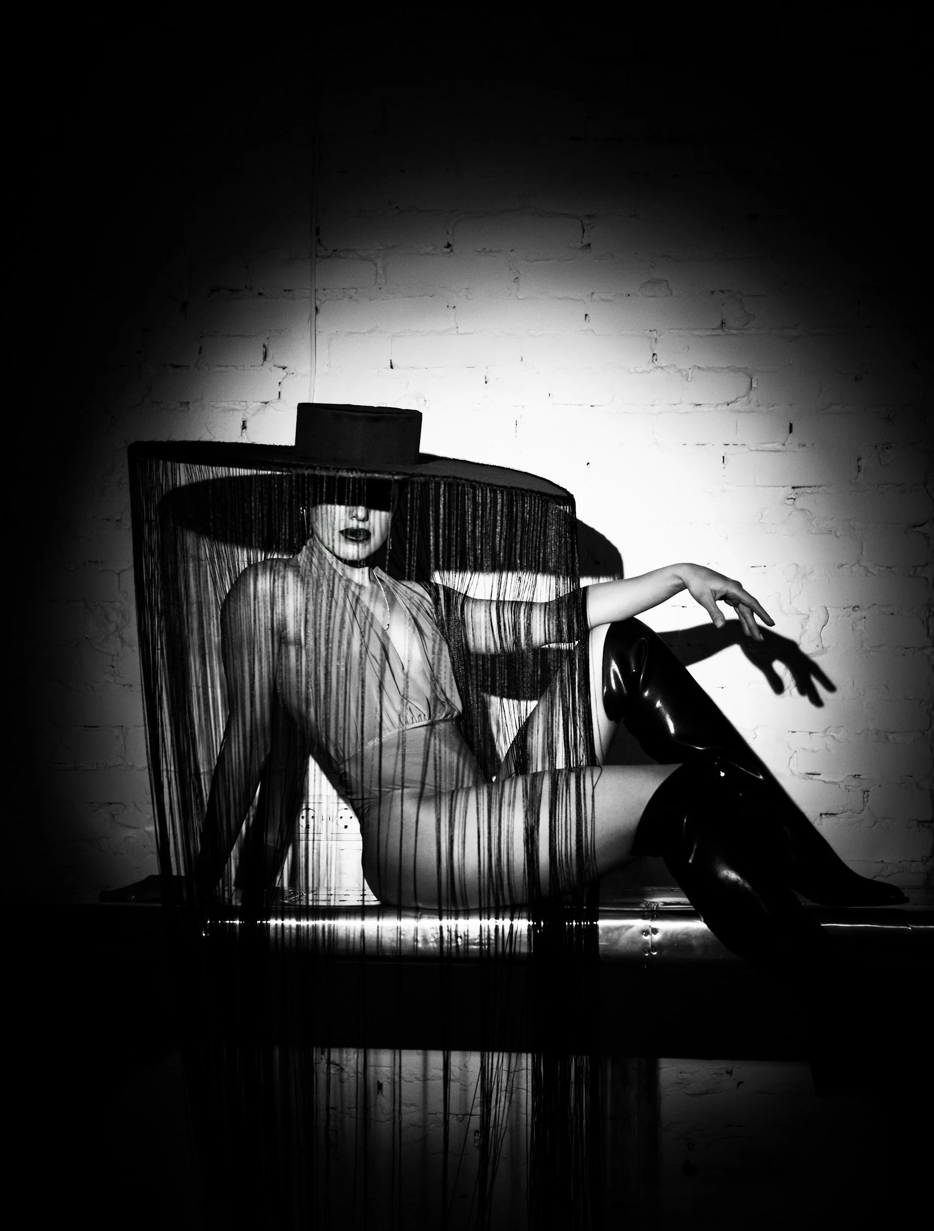 A woman wearing a hat and sheer clothing, sitting in a chair, with a fringe covering her eyes, against a brick wall background, in a black and white photograph.