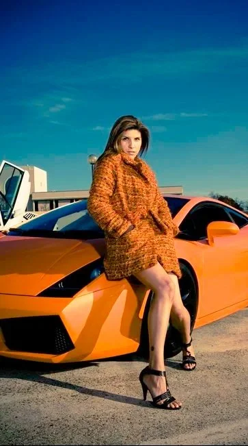 A woman in an orange coat and black high heels leaning against an orange sports car outdoors under a clear blue sky.