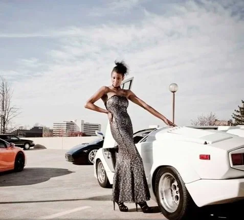 A woman in a long leopard print dress standing next to a white sports car in a parking lot.
