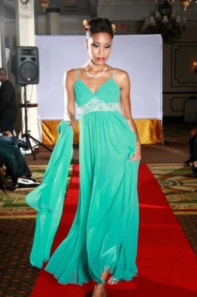 A woman walking on a red carpet wearing a flowing turquoise dress with a lace waist, holding part of her dress in one hand at an indoor event.