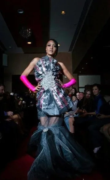 Model walking on runway in an elaborate dress with layered fabric and a long tulle train, wearing bright pink gloves, at a fashion show.
