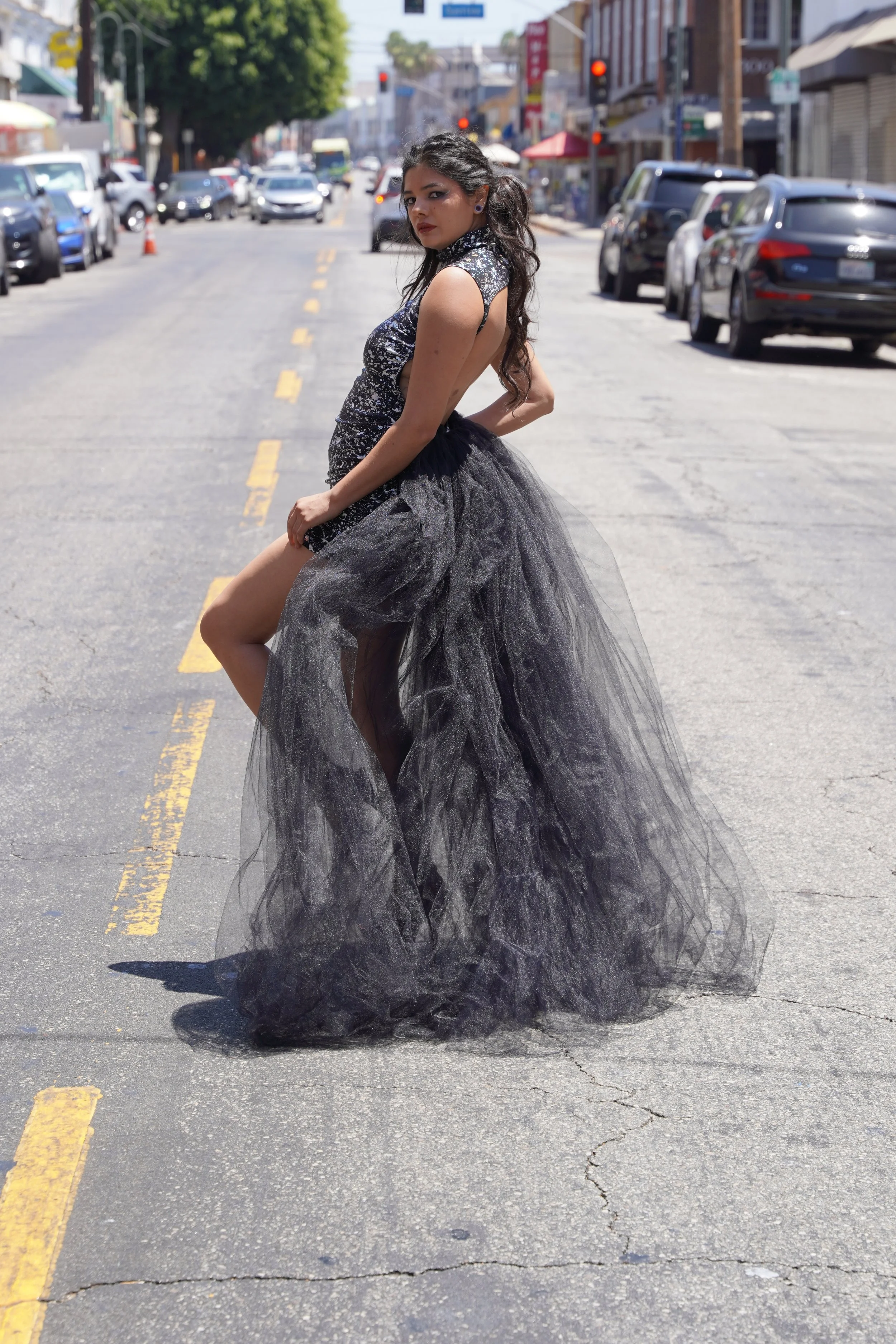 A woman in a glamorous black and silver dress with a tulle train stands in the middle of a city street, with cars parked along the side and buildings in the background.