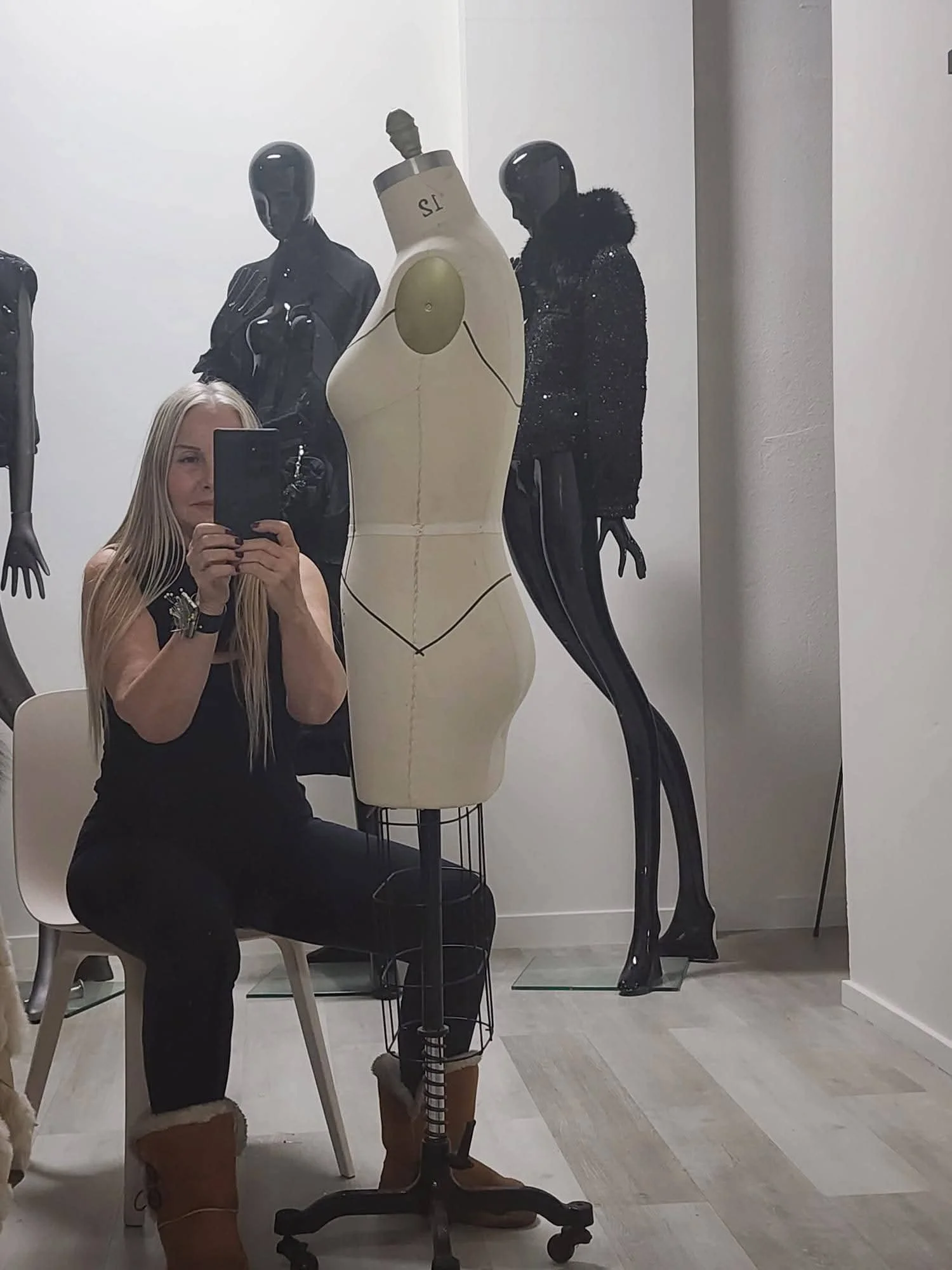 A woman taking a mirror selfie in a store fitting room. Behind her are mannequins, one nude and one dressed in black, with a white wall and gray flooring.
