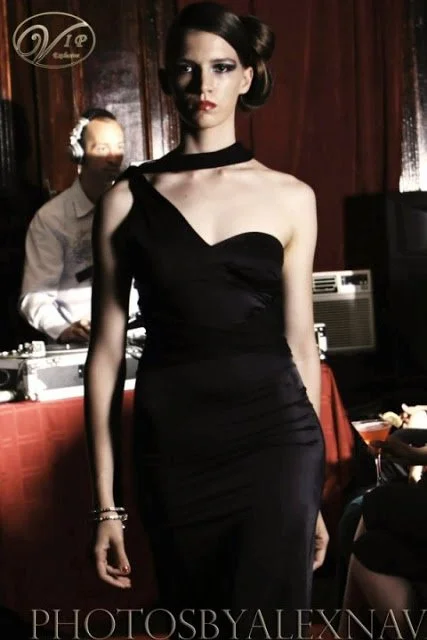 A woman with dark hair styled in an elegant updo wearing a black, asymmetrical, one-shoulder evening gown at an indoor event, with a DJ in the background.