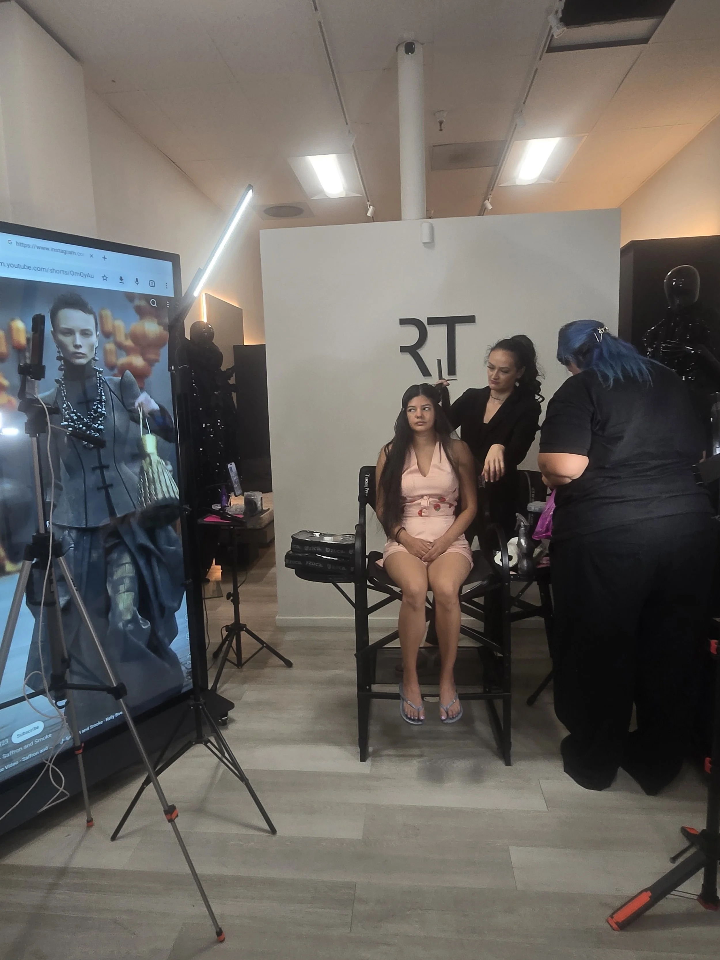 A photoshoot in progress with a woman sitting on a chair, being styled by two stylist women. The woman wears a pink dress with a cherry pattern, decorated earrings, and silver high heels. One stylist with dark hair is fixing her hair, while the other