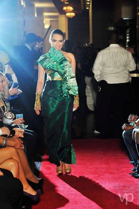 A female model walks down the runway in an elegant green dress with floral ruffle details, gold gloves, and high heels at a fashion show. Audience members are seated on either side, and a photo is being taken.