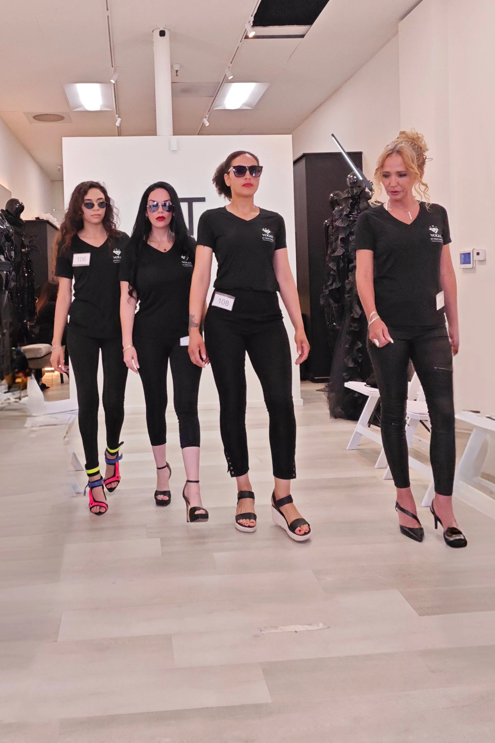 Four women walking in a fashion show, wearing black T-shirts and black pants, with different stylish shoes and sunglasses.