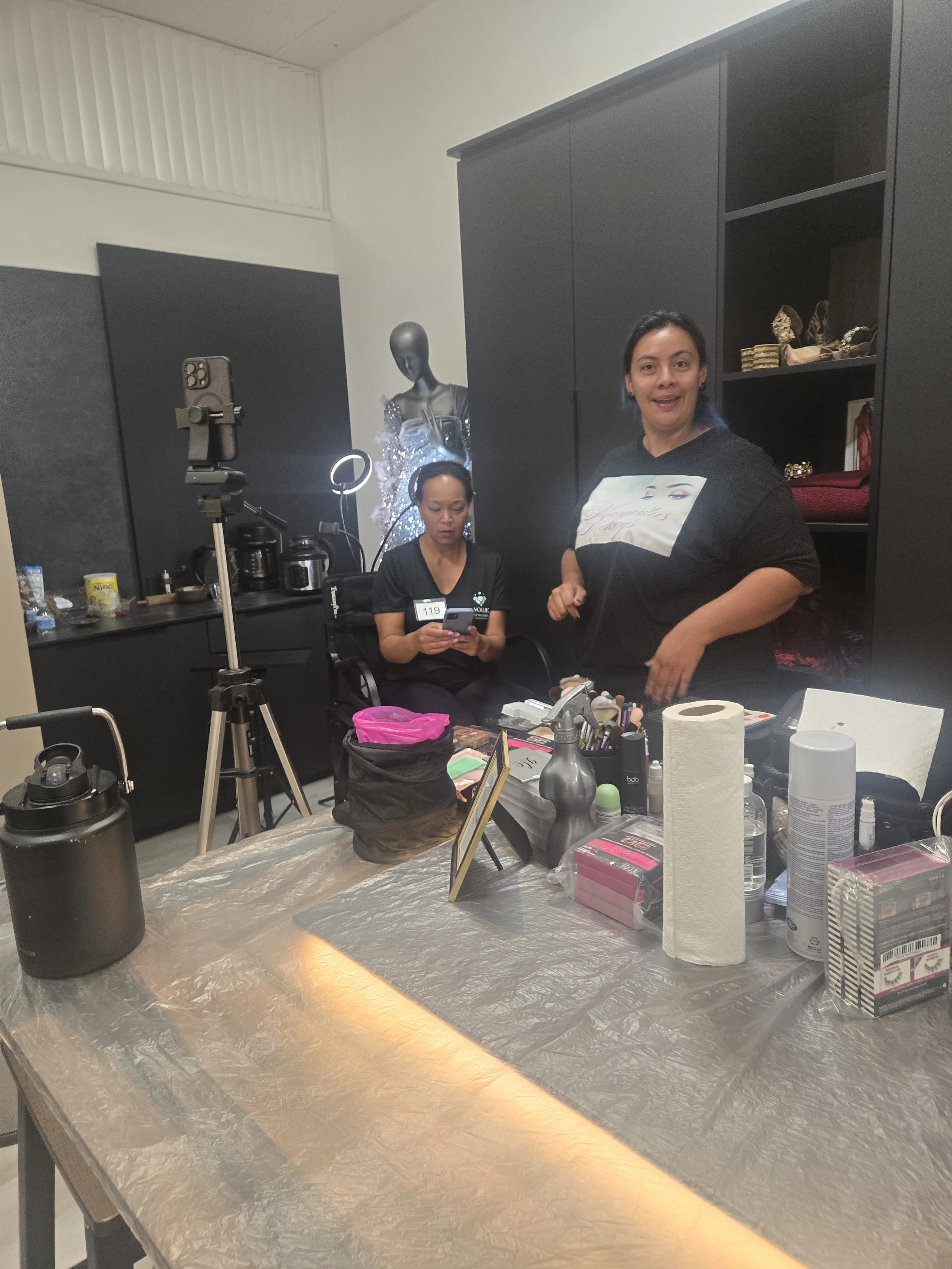 A room with two women working at a makeup or beauty station. One woman is standing and smiling at the camera, the other is sitting and looking at a phone. There are makeup supplies and tools on the table, and a mannequin dressed in a glittery outfit 