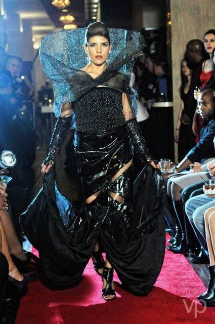 A model walking down a runway at a fashion show, wearing a black and blue avant-garde outfit with large, dramatic shoulder pieces, shiny black pants, and high heels.
