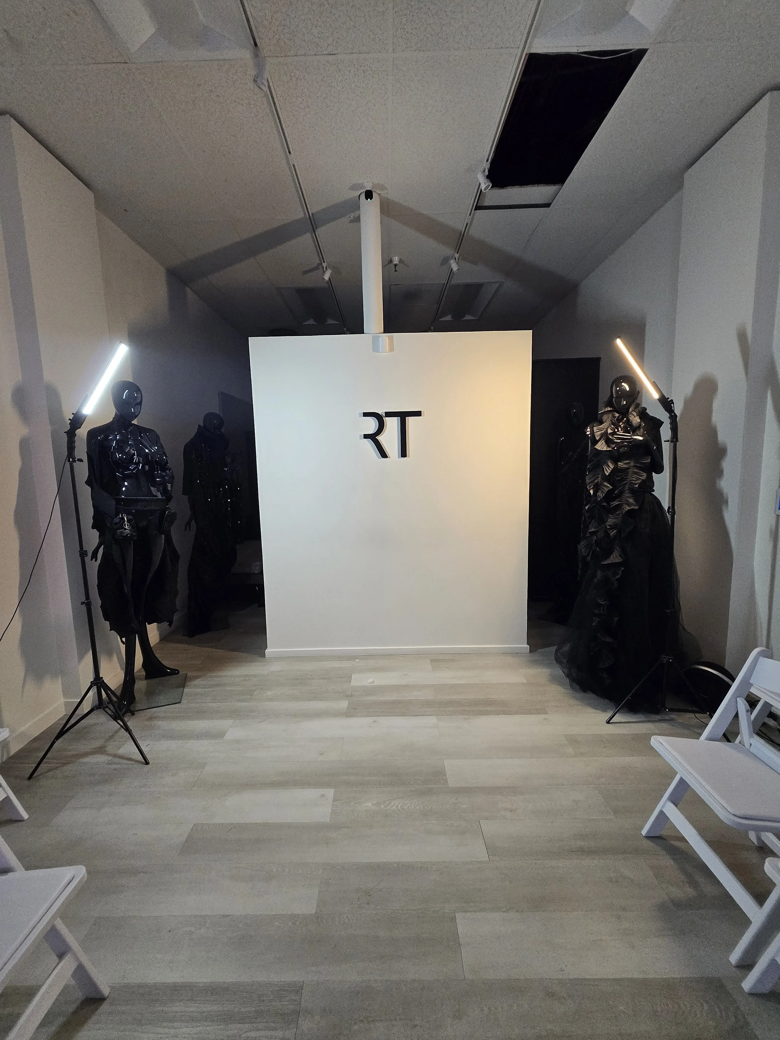 Interior of a studio or showroom with mannequins dressed in black clothing, a white display board with 'RT' on it, and professional lighting equipment.