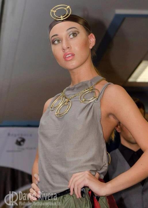 A woman modeling a fashion outfit with large jewelry, wearing a gray sleeveless top, posing confidently at an indoor event.