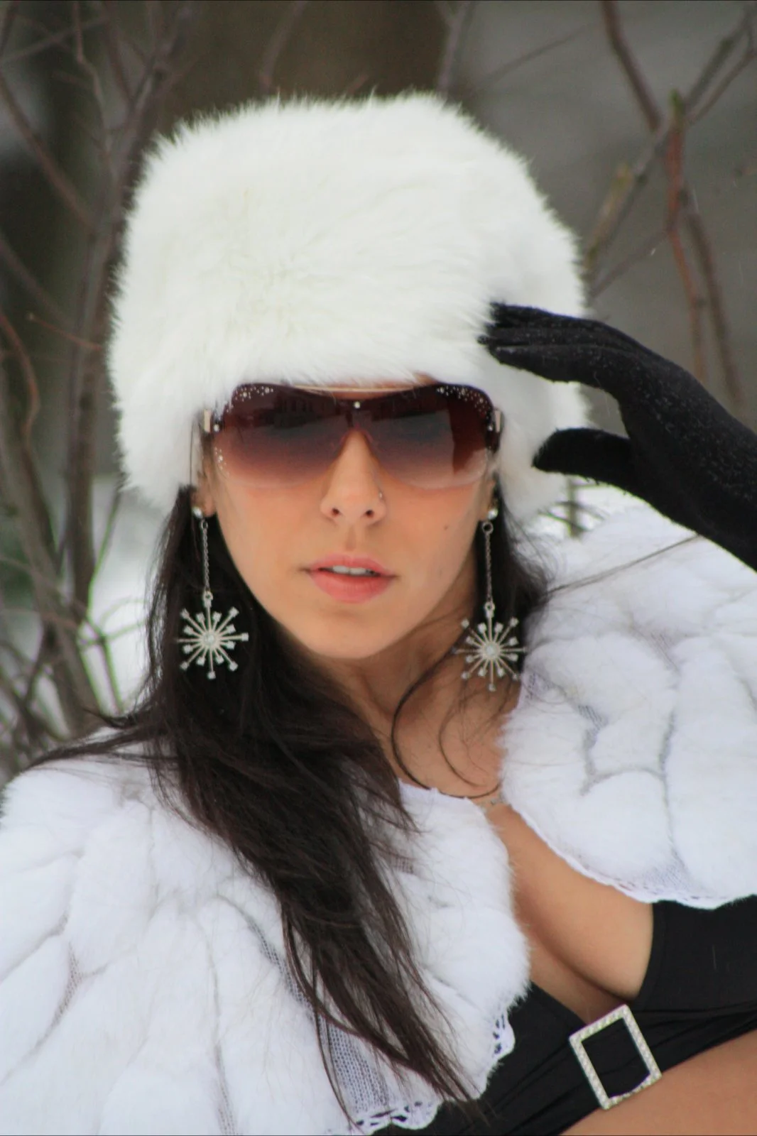 Woman wearing sunglasses, large white furry hat, black gloves, and earrings with snowflake design in a winter outdoor setting.