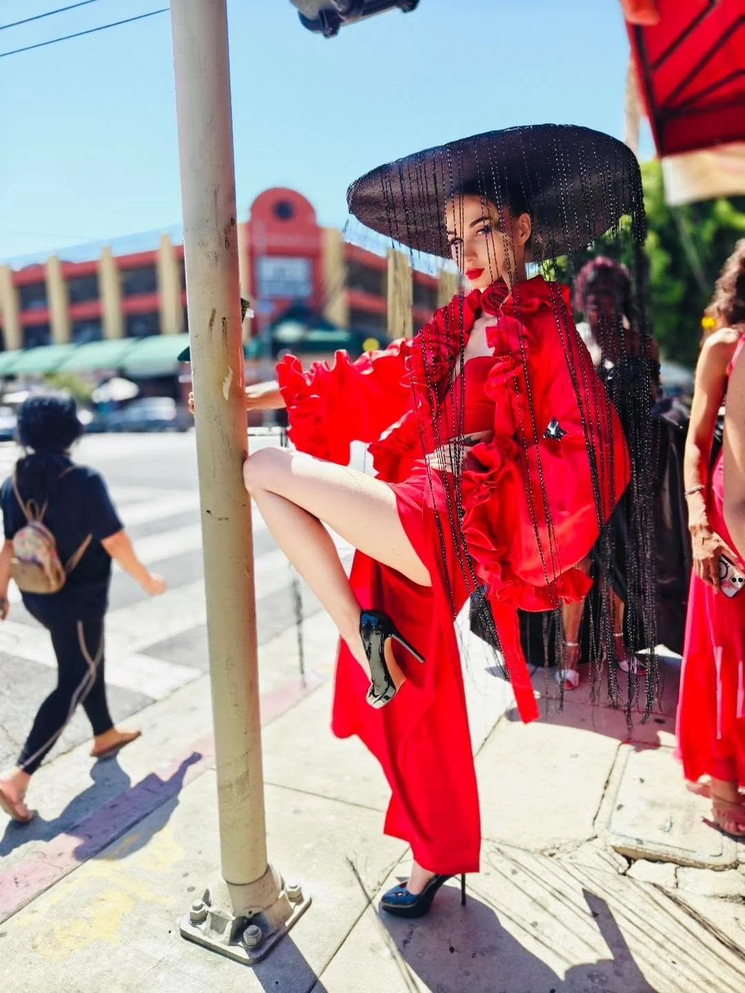 Woman in a red dress and large black hat with veil standing on a sidewalk during daytime.