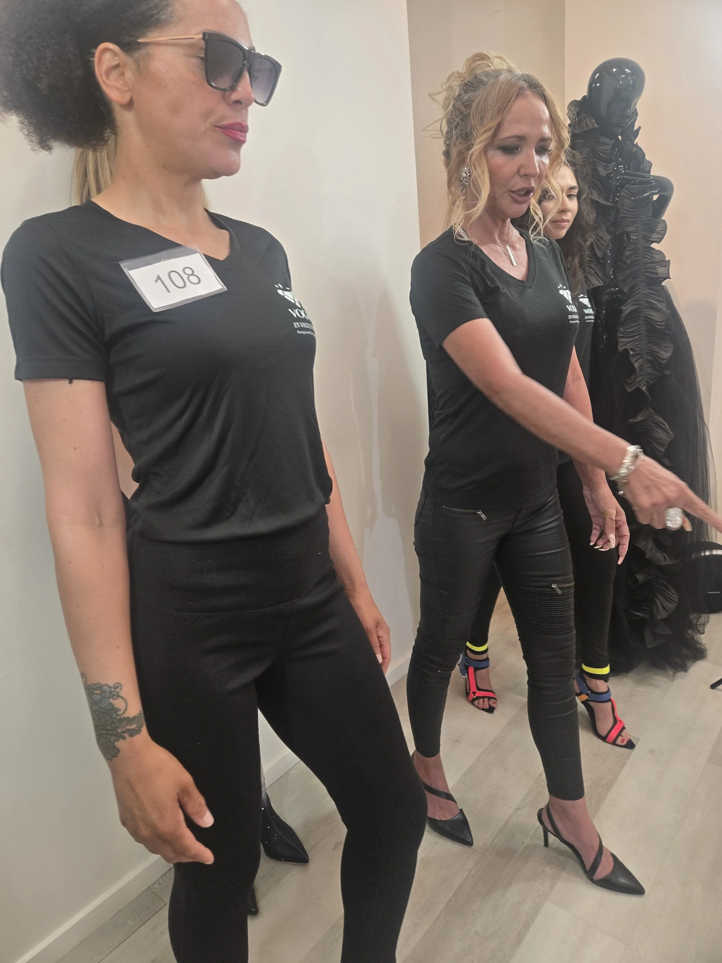 Four women standing in a line against a wall, wearing black shirts, black pants, and high heels, with some accessorized with jewelry, at a fashion event or show.
