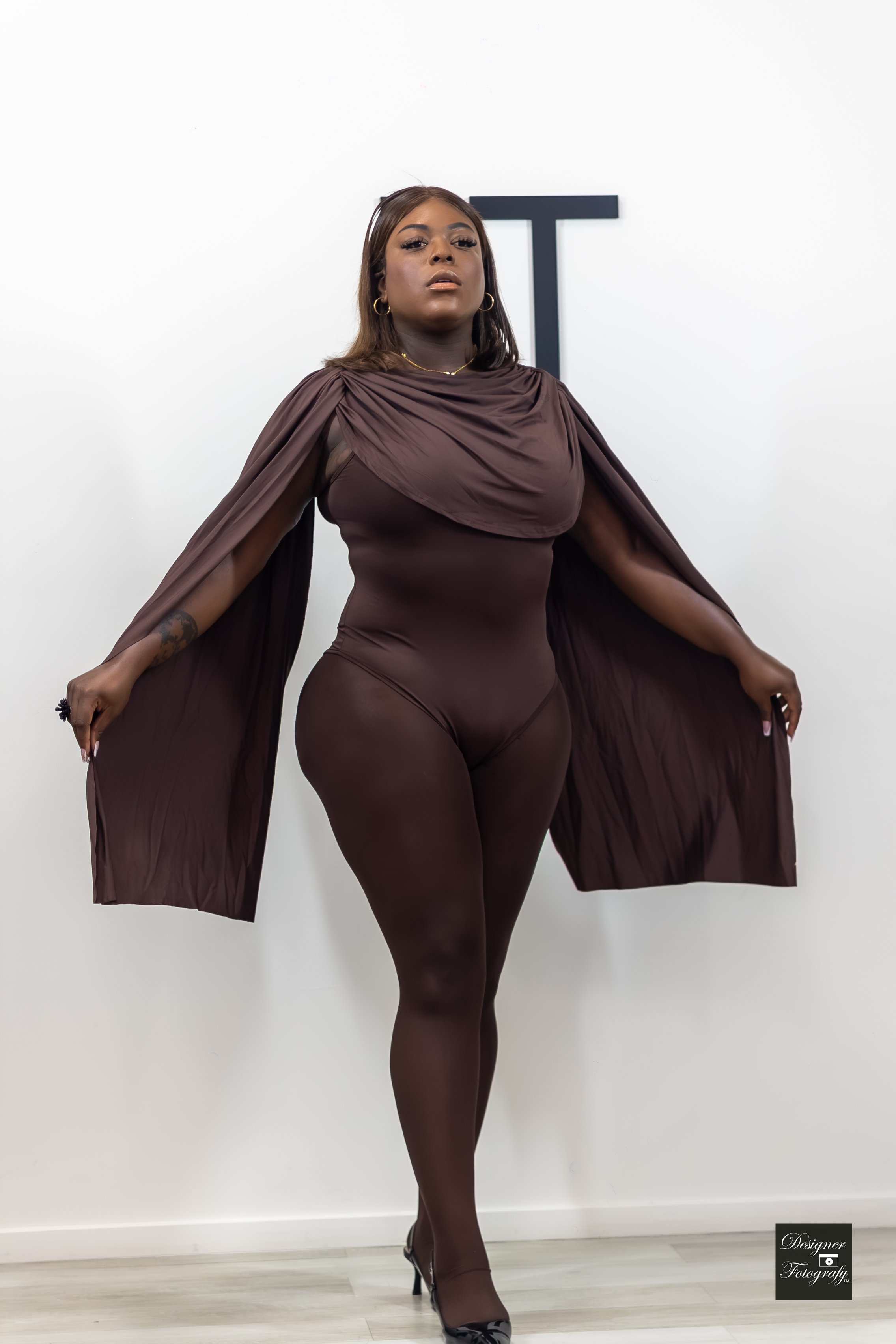 A woman with dark skin in a brown outfit stands against a white wall. She has shoulder-length brown hair, gold hoop earrings, and is wearing high heels. Her pose is confident with her arms extended to the sides, and she is looking directly at the cam