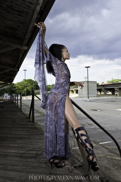 A woman in a purple lace dress standing on a wooden platform outdoors, posing with one arm raised and her leg extended, against a cloudy sky and urban background.