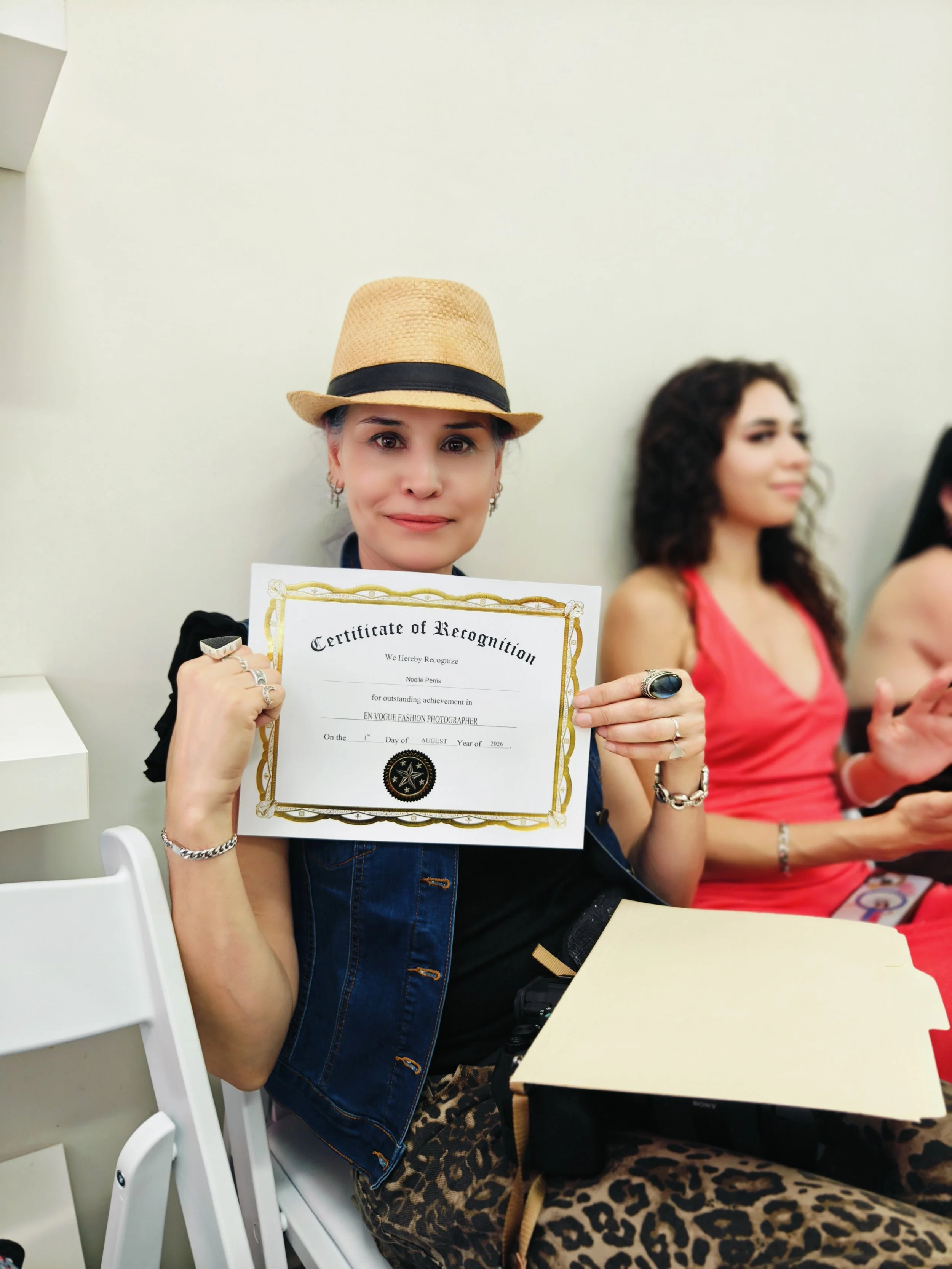 Woman wearing a straw fedora hat and leopard print pants holding a certificate of recognition, sitting at a table with other people in the background.