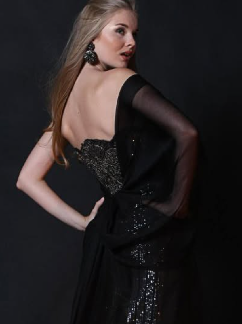 A woman with long blond hair wearing a black dress with sparkly details, large black earrings, and sheer black sleeves, posing against a black background.