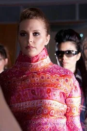 A woman wearing a colorful, patterned dress with a high neckline, standing in a crowd with people in the background, including a woman with sunglasses.
