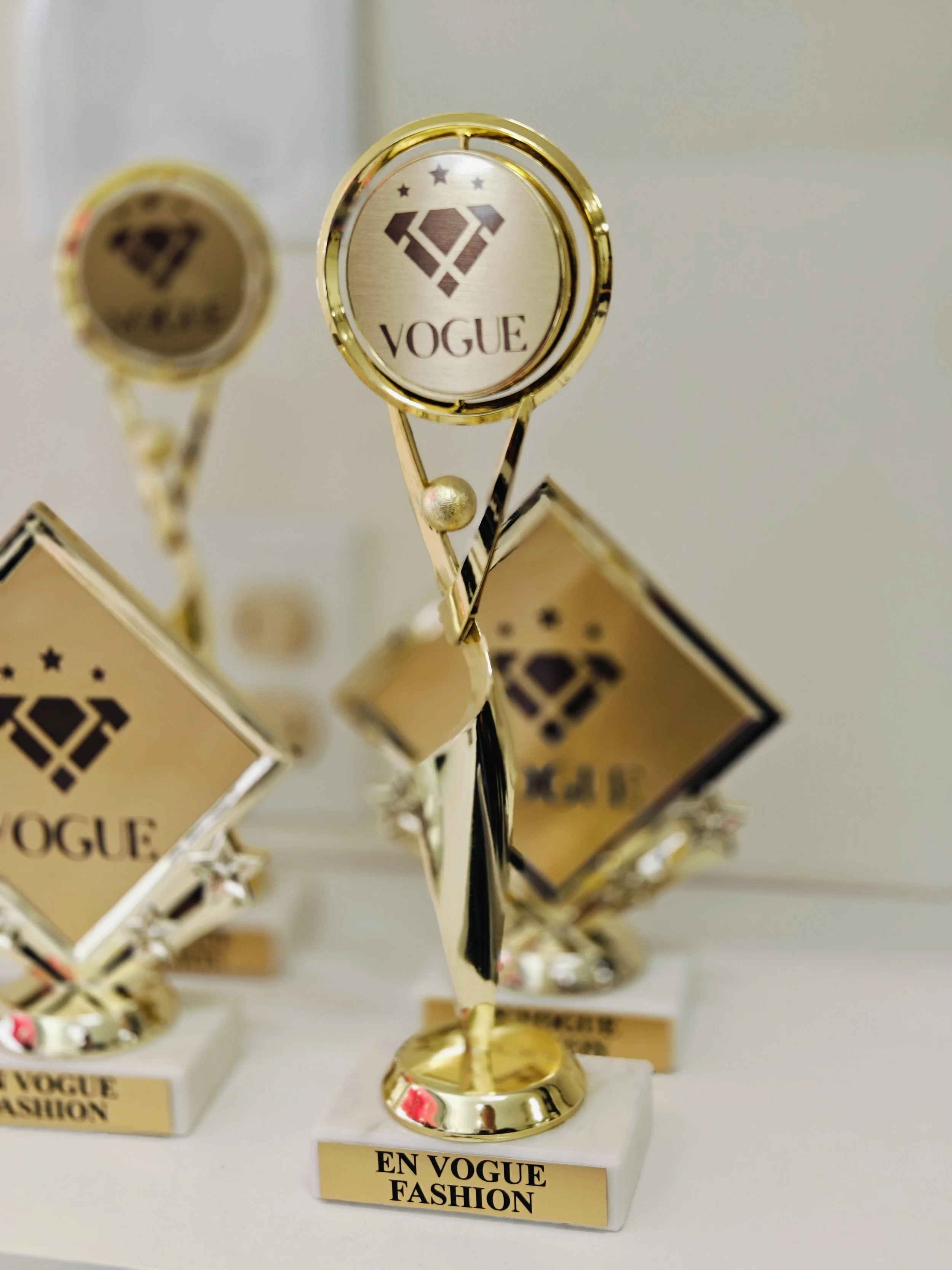 A gold-colored trophy with a circular top featuring a diamond-shaped logo and the word 'VOGUE' underneath. The trophy has a star engravings above the logo, a slender stem, and a round base. It is displayed on a white surface with a label at the front