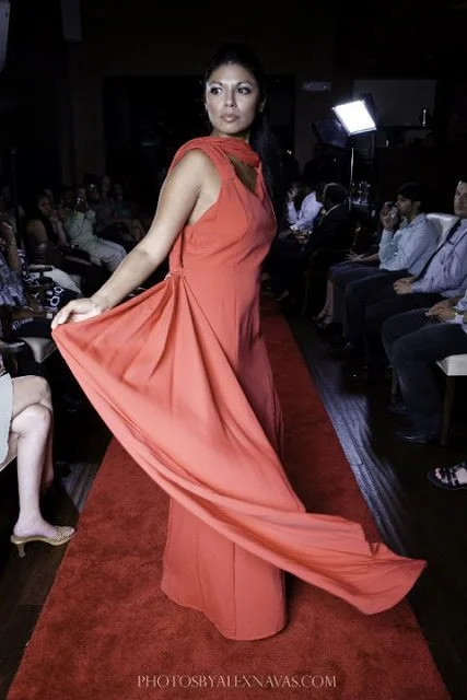 A woman in a long, flowing coral-colored dress stands on a red carpet, holding out her dress with one hand.