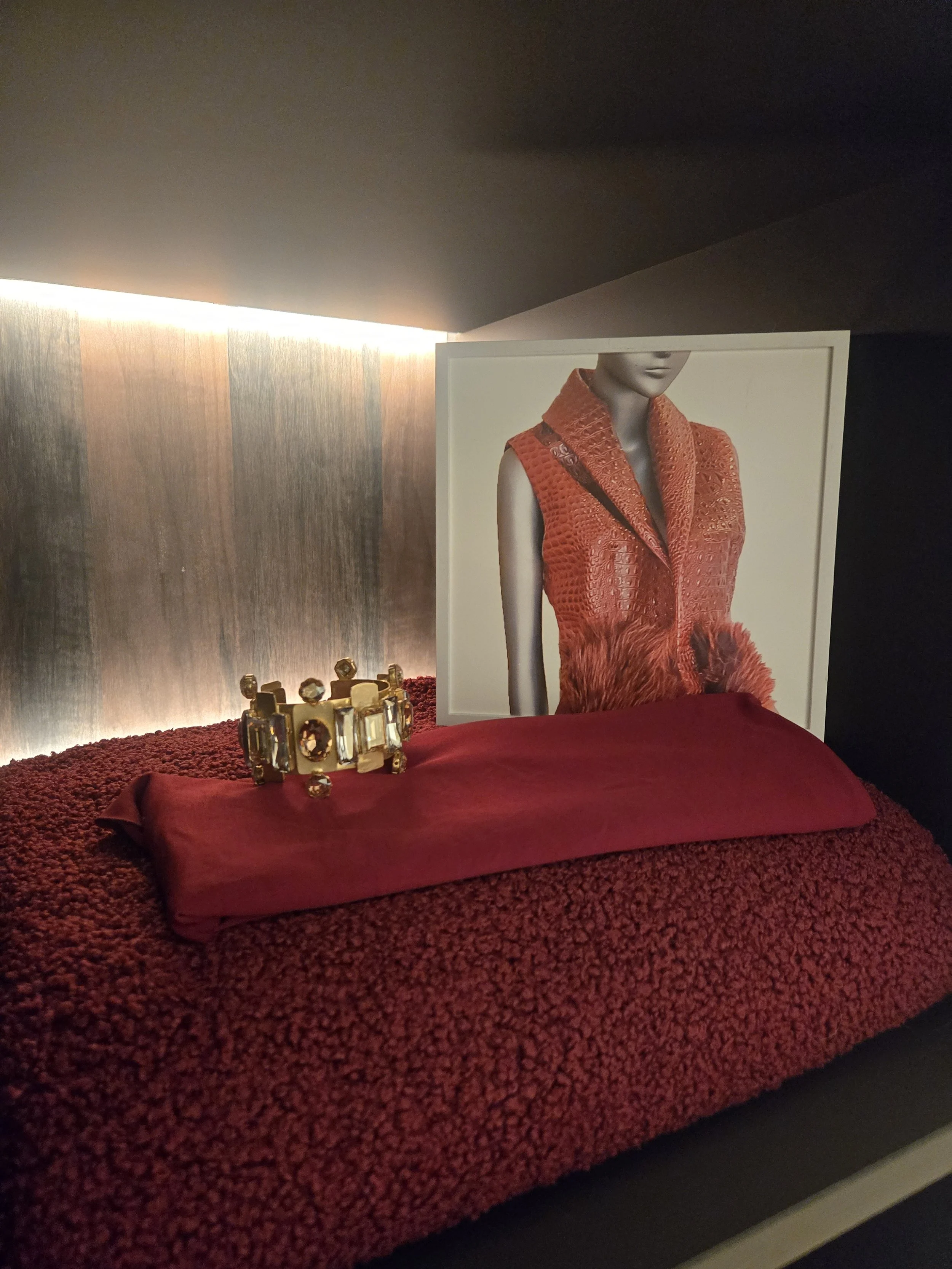 Decorative display with a framed fashion photograph, gold jewelry bracelet with large gemstones, and a red textured fabric on a dark surface.