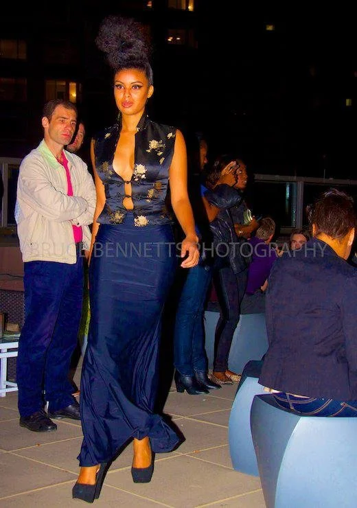 A woman in a long navy blue dress with a lace top and cut-out details, standing on an outdoor terrace at night. Several other people are in the background, some sitting and some standing.