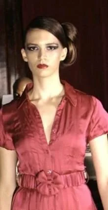 Young woman with dark hair styled in a side bun wearing a red satin blouse with a tied waist, standing indoors with a serious expression.