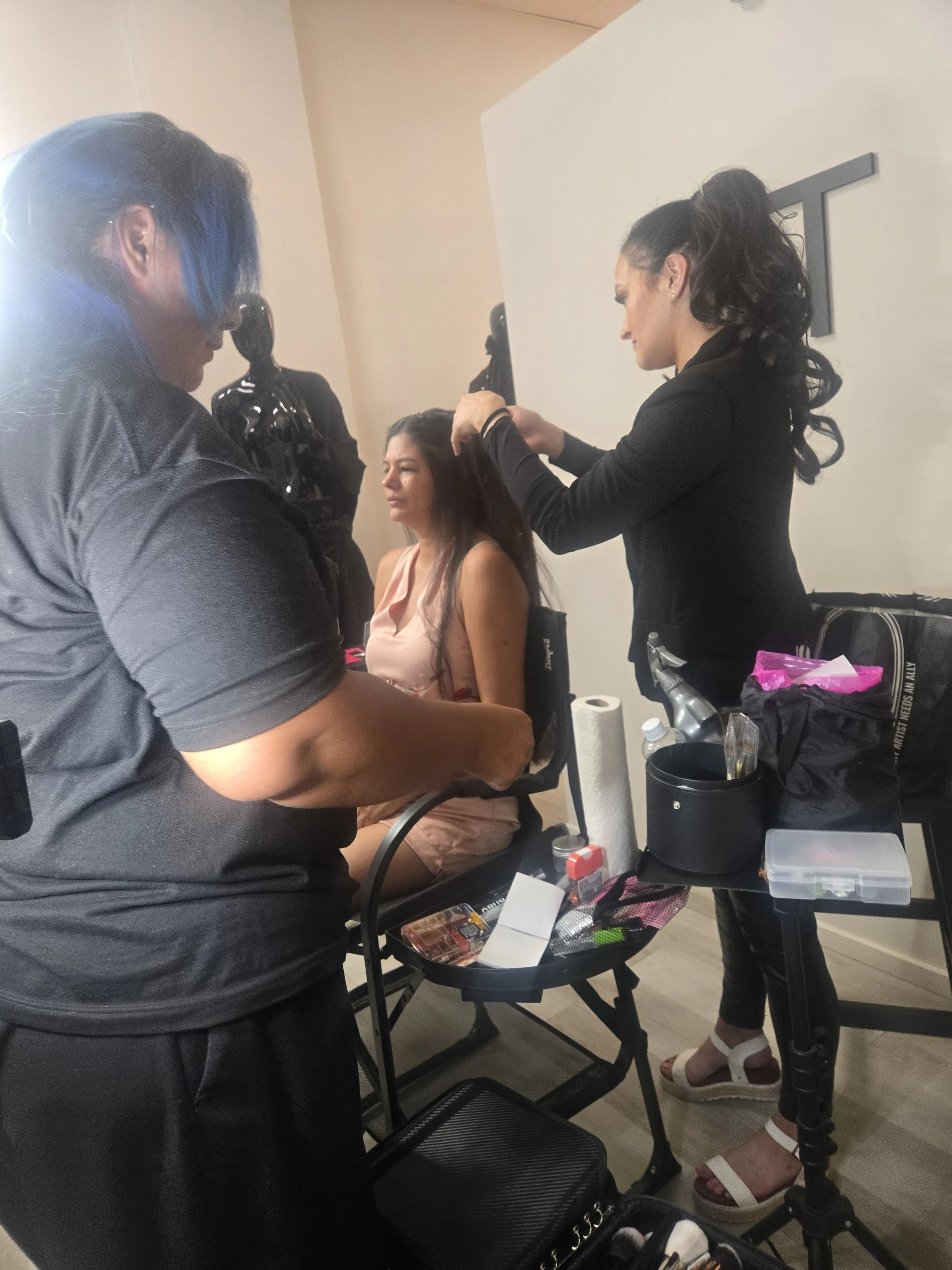A woman with dark hair and tan skin sitting in a makeup chair while another woman styles her hair. Various makeup tools and supplies are on a cart nearby.