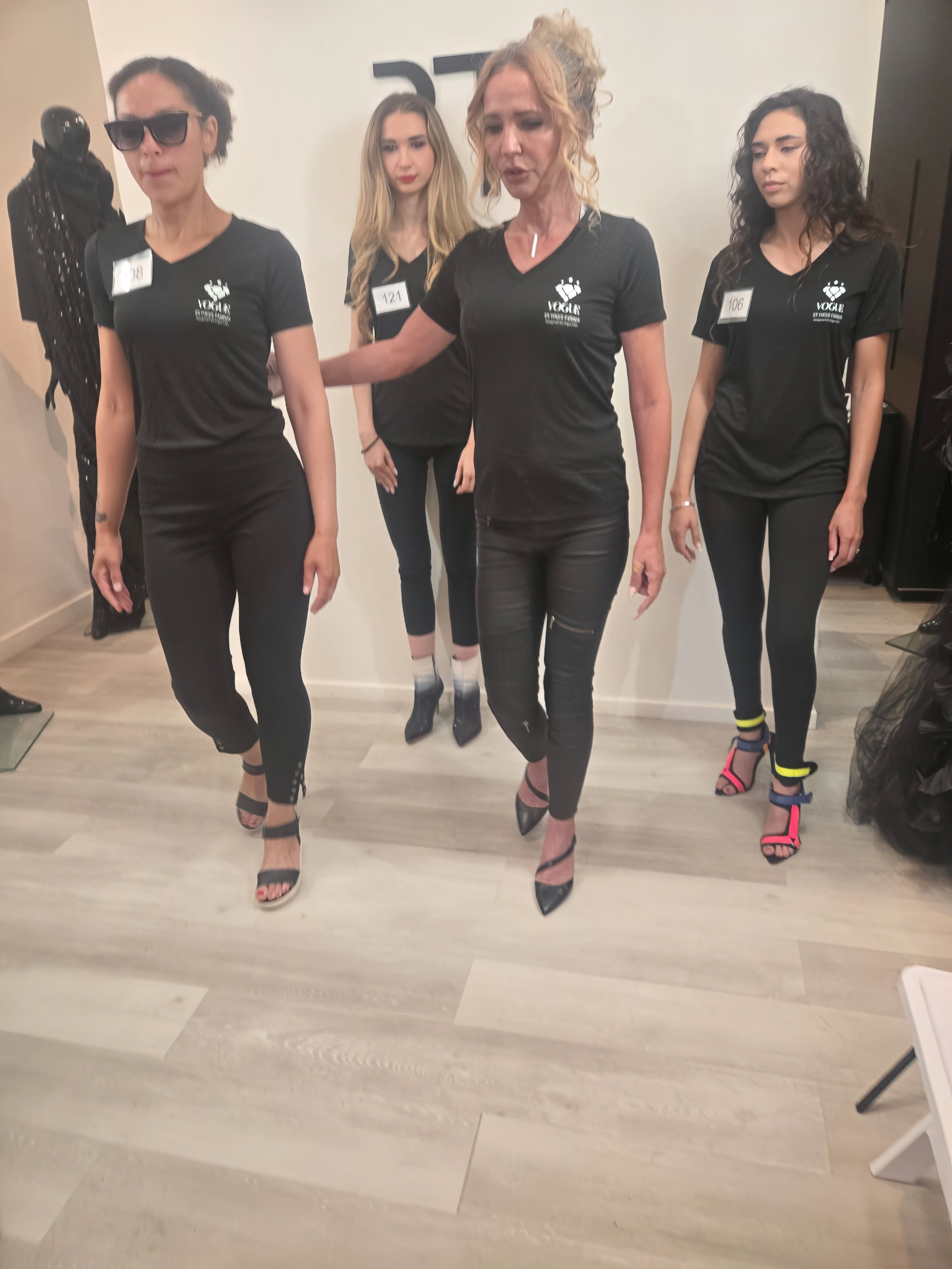 Four mannequins dressed in black outfits in a boutique or showroom, with one mannequin wearing sunglasses and a snake-skin patterned outfit, and another in high-heeled shoes with colorful straps.