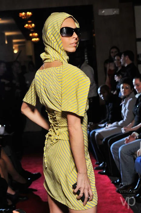 A woman in a yellow and white striped dress with a matching hood, large sunglasses, and holding a phone, walking on a runway during a fashion show with seated audience in the background.