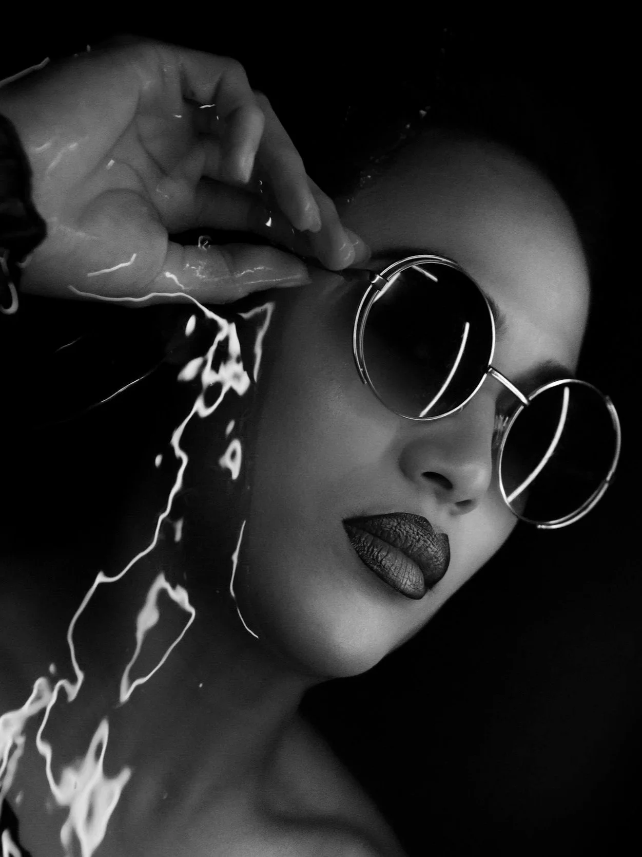 A black and white photo of a woman wearing round sunglasses and dark lipstick, holding a glass jar near her face with water splashing out.