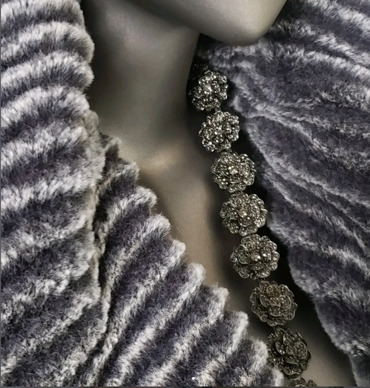 A close-up of a jewelry mannequin with a grey velvet scarf, a furry grey shawl, and a sparkly flower-shaped rhinestone necklace.