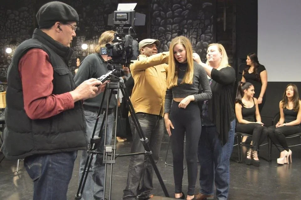 People on a film set, with a camera crew and actors, preparing for a scene. An actress with red hair is being helped with her wardrobe by two women, while others sit or stand in the background.