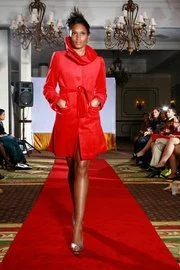 A woman walking down a runway wearing a red coat and dress, with a chandelier and seated audience in the background.