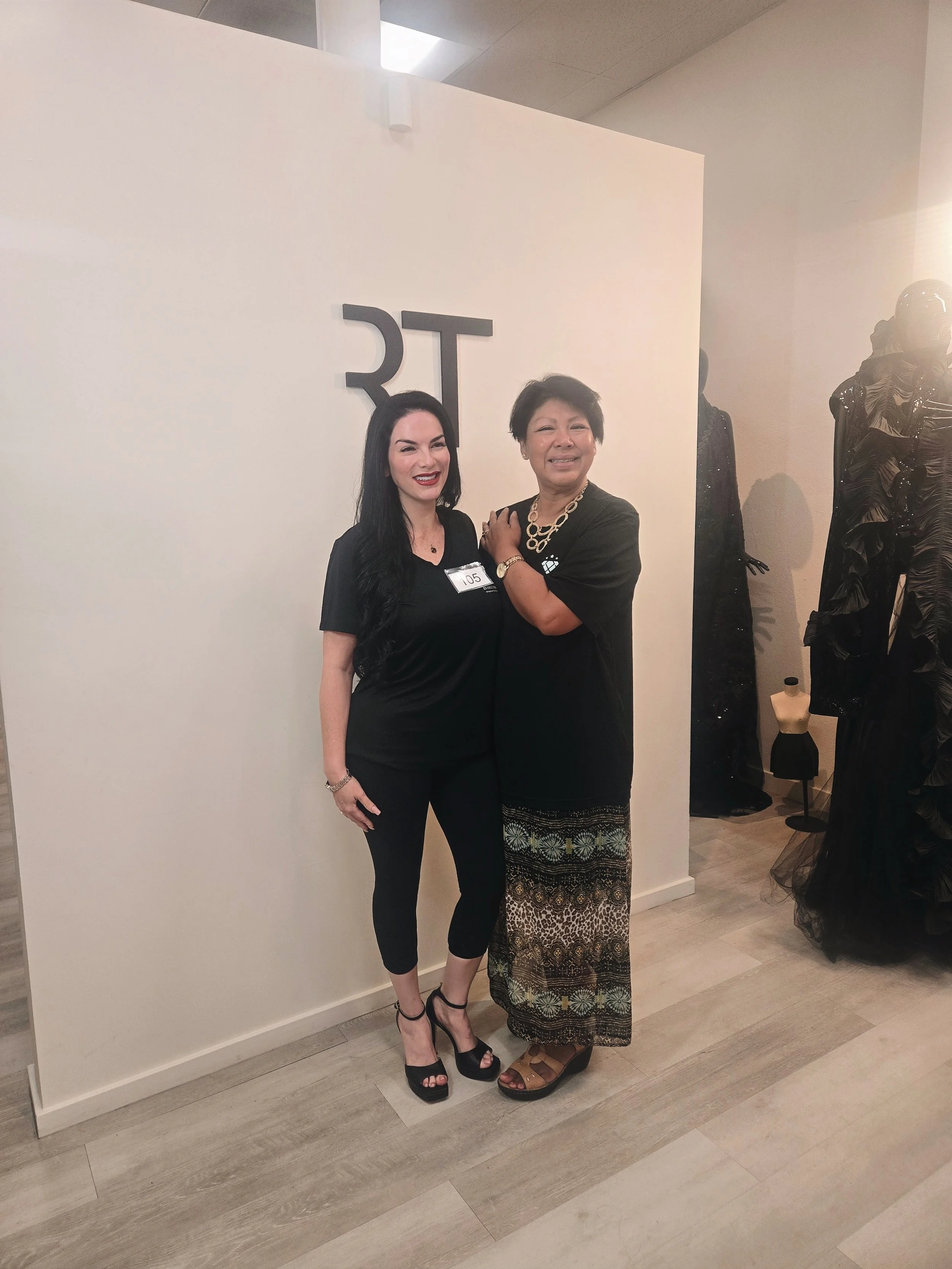 Two women are standing together smiling in an art gallery or fashion store, one wearing a black dress and high heels, the other in a black top and a long patterned skirt, with mannequin displays in the background.