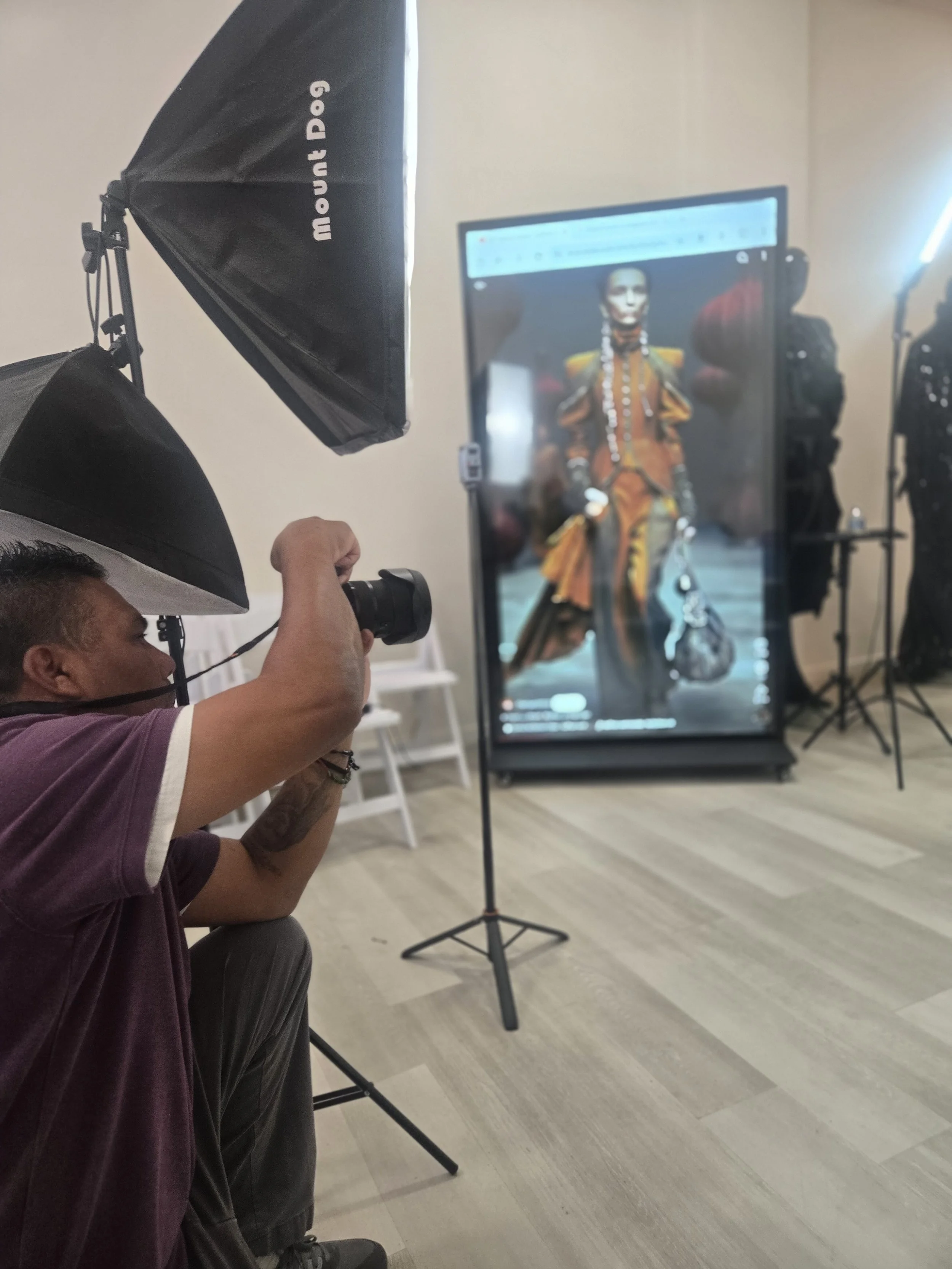 A person crouching in front of a photography setup with a camera in hand, capturing a model on a large monitor that shows the model in an elaborate, colorful costume. The setup includes studio lights and a black backdrop.