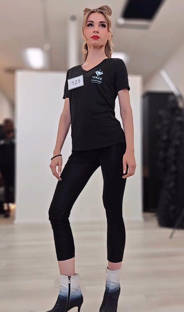 A young woman standing confidently in a fashion show setting. She is wearing a black t-shirt with a logo and a name tag, black shiny leggings, and high-heeled boots with a gradient design. She has her hair styled in two buns and is looking upward.