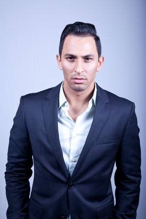 A young man in a navy suit and white shirt standing against a plain light gray background.