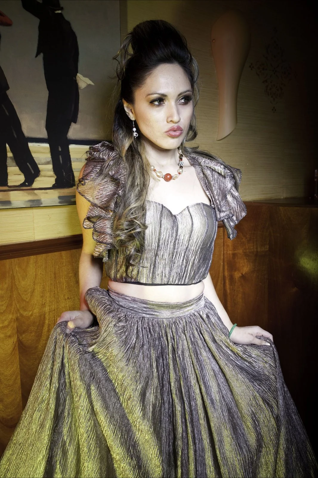 A woman with long wavy hair wearing a metallic colored ruffled top and matching wide skirt, holding the skirt with both hands, standing indoors with wooden wall and artwork in the background.