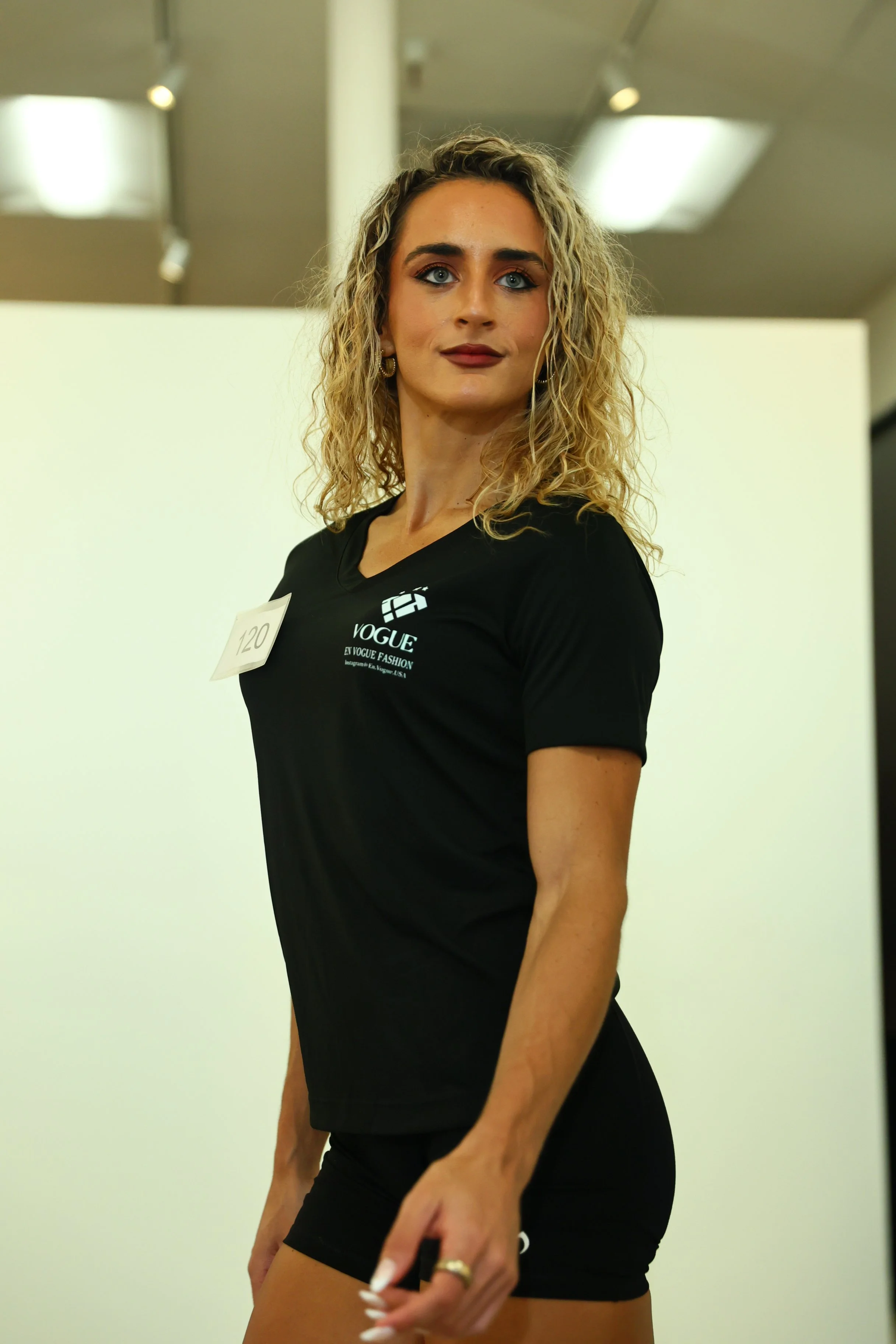 Young woman with curly blonde hair and blue eyes, wearing black clothing and jewelry, standing indoors with a neutral expression.