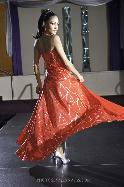 A woman in a red strapless ball gown with a train, standing on a stage with purple curtains and mirrors in the background.