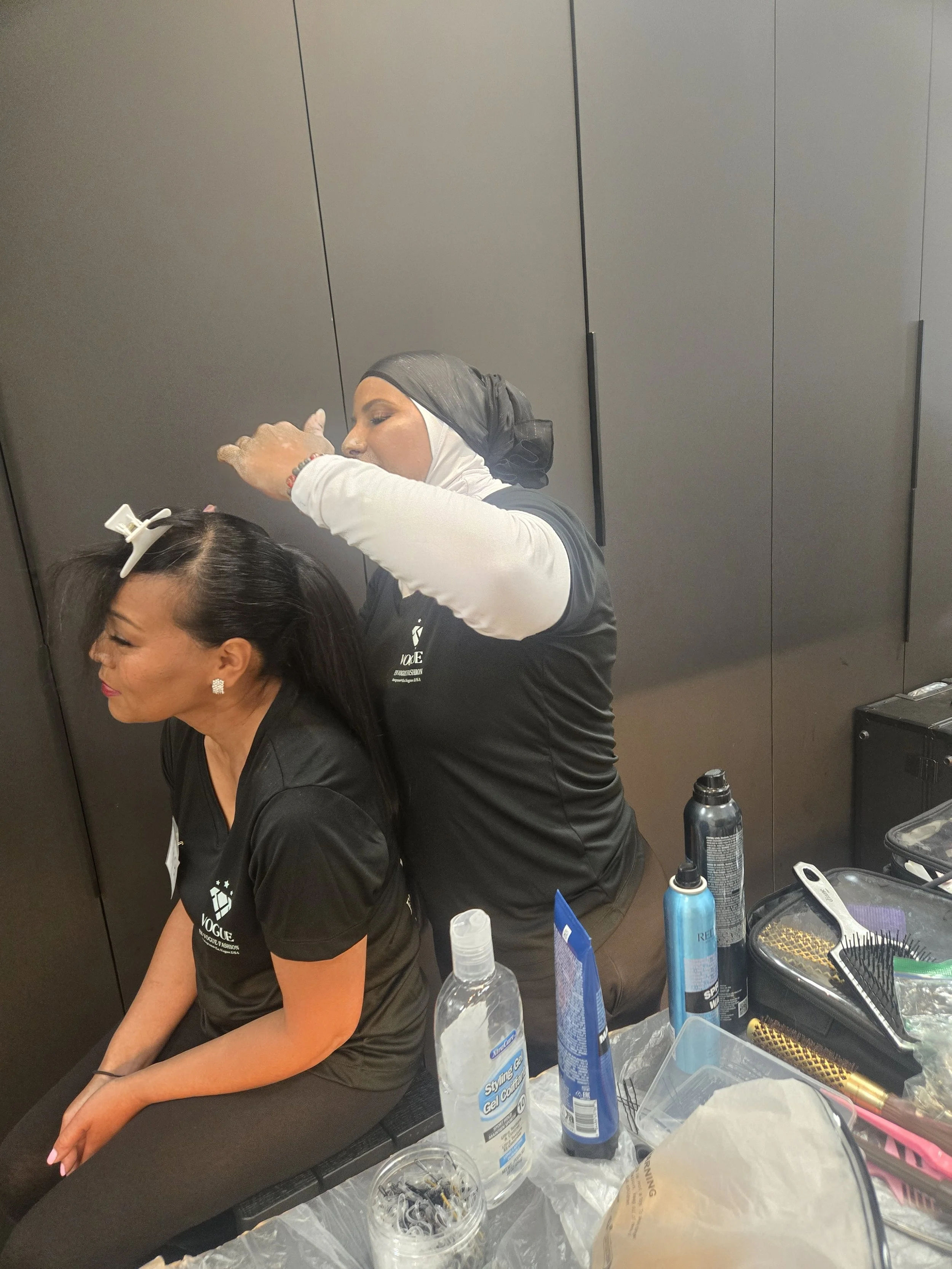A woman with long dark hair, seated, getting her hair styled by a stylist in a dressing room. The stylist wears a black shirt and a gray headscarf. The table in front of them has hair styling products, tools, and accessories.