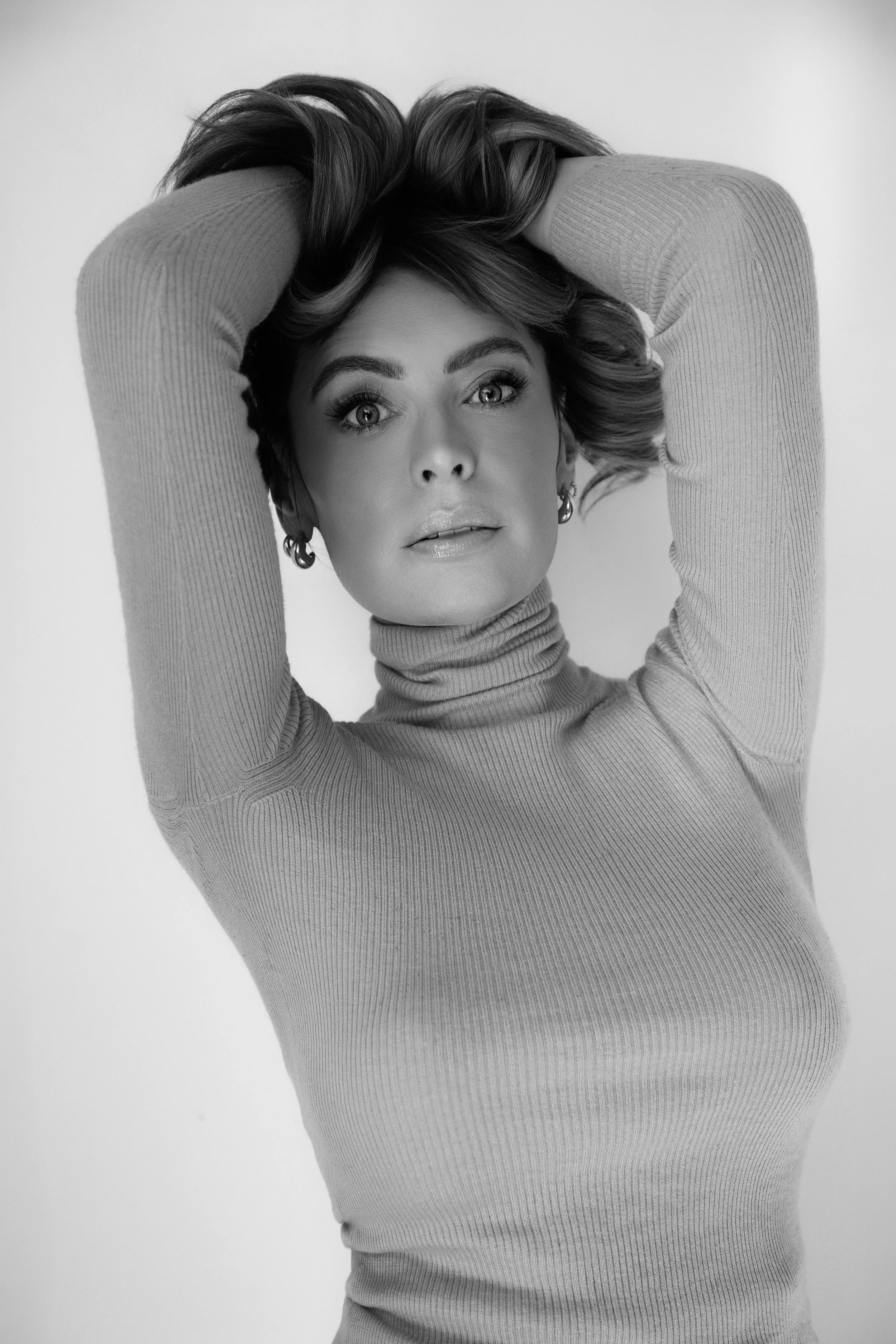 Black and white portrait of a woman with big eyes and styled hair, wearing a turtleneck sweater with her hands in her hair.
