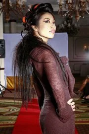 A woman with long black hair in an elegant updo, wearing a fitted maroon dress, standing sideways in a decorated indoor setting with a red carpet.