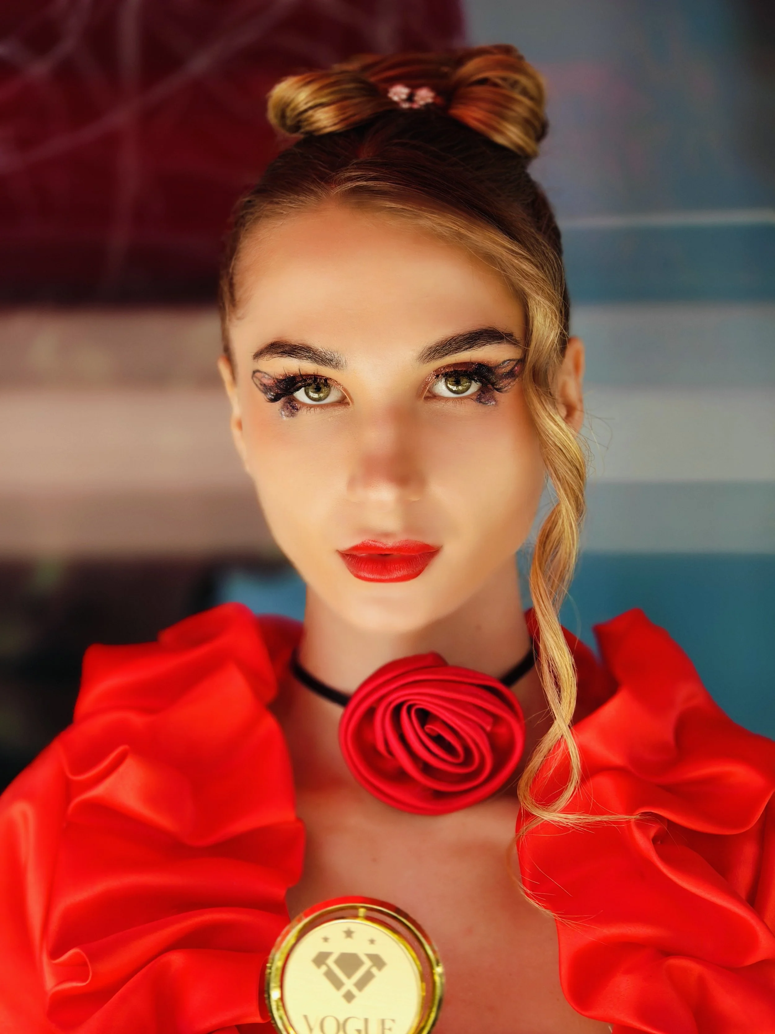 Close-up of a woman with platinum blonde hair styled in a voluminous updo with a braid, green eyes with makeup, red lipstick, wearing a red dress or jacket with ruffled sleeves, and a large red rose necklace.